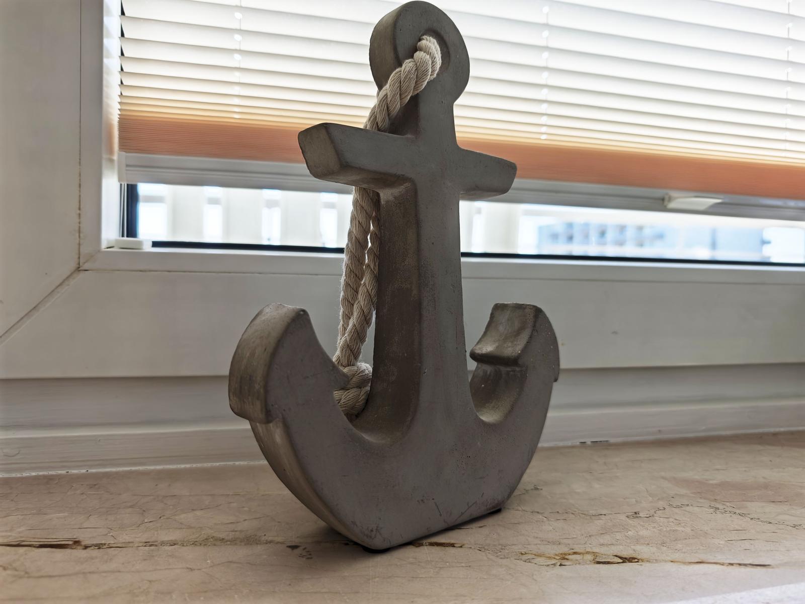 Ein grauer Anker mit Seil steht auf einer Steinplatte vor einem Fenster.