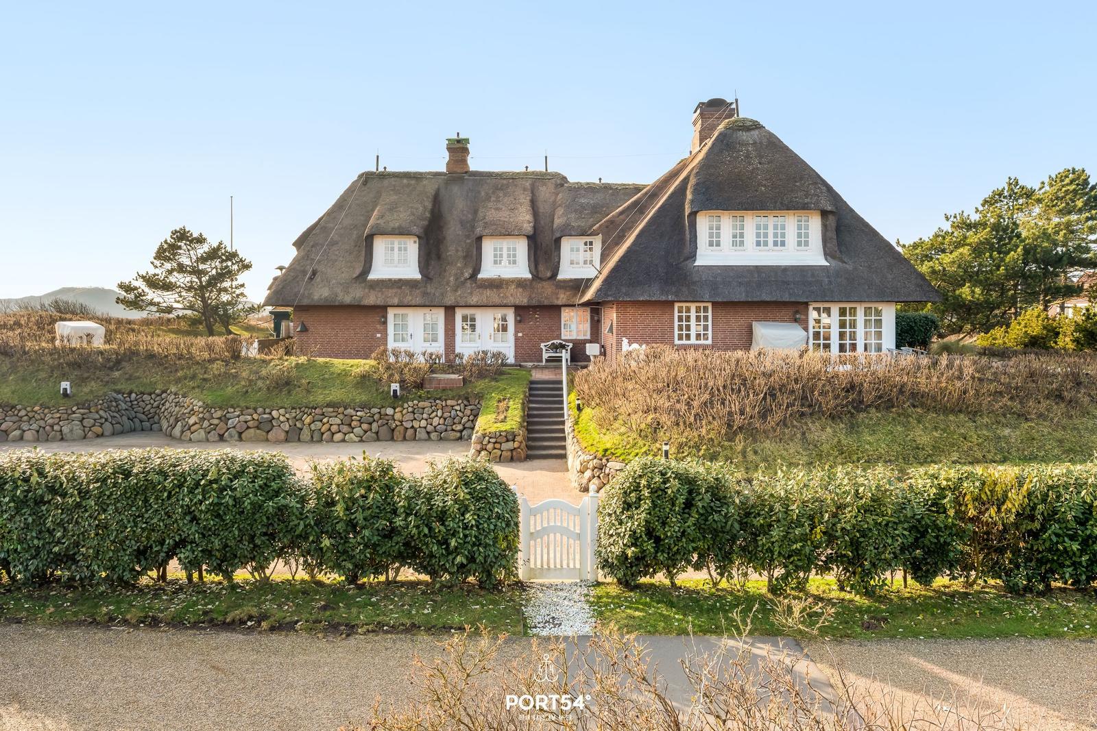Haus mit Strohdach, weißen Fenstern und Steinmauern. Eingangstreppe und Hecke sichtbar.