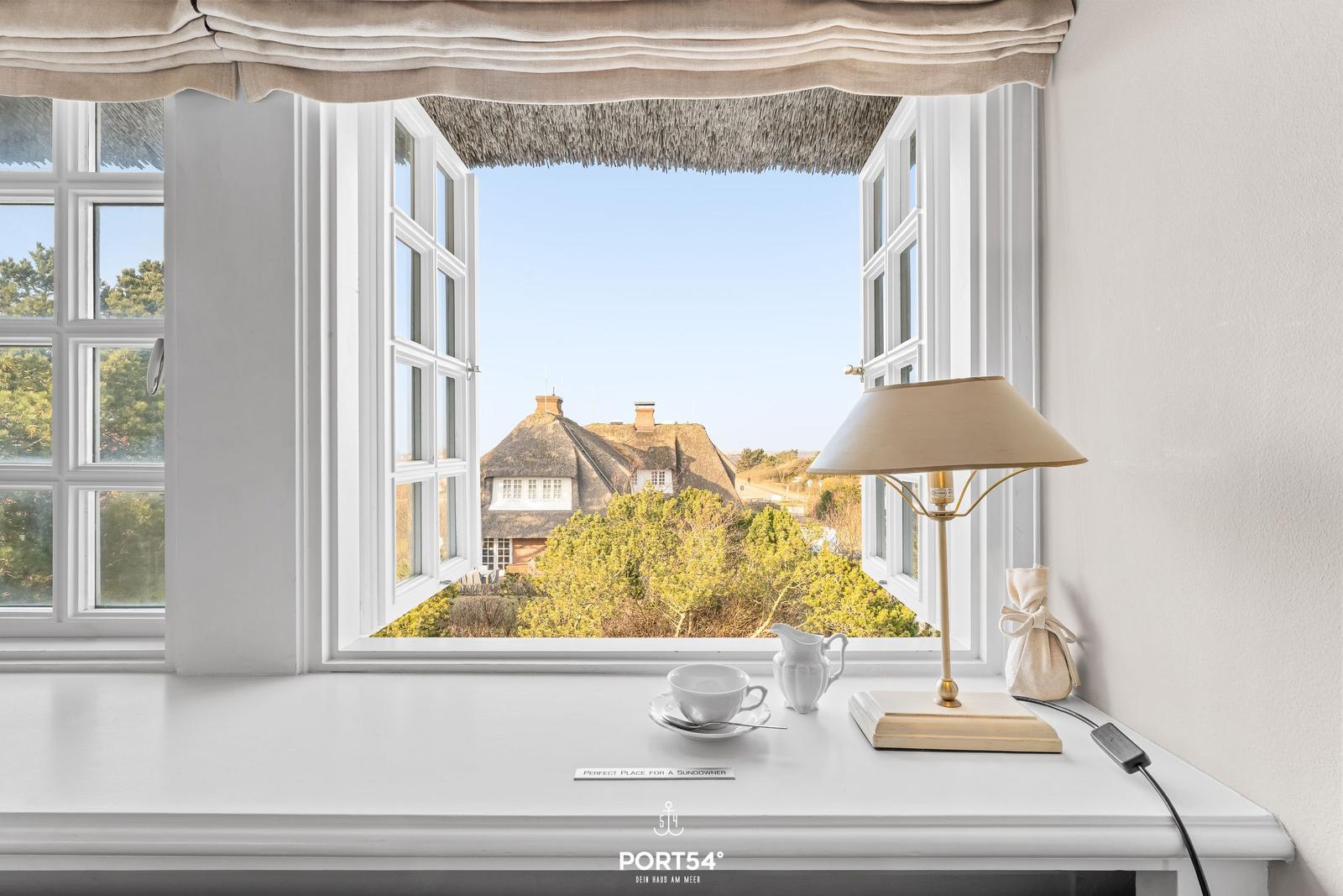 Weißer Fensterbalken mit Tasse, Lampe und Blick auf strohgedecktes Haus.