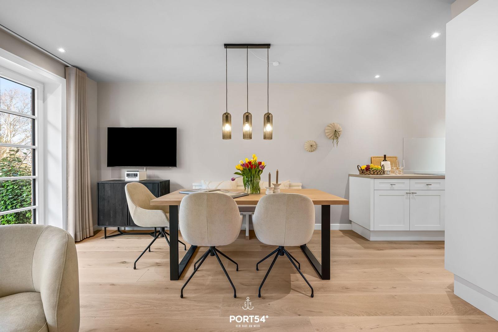 Esszimmer mit Holztisch, vier Stühlen und moderner Beleuchtung. Küche mit weißen Schränken im Hintergrund.