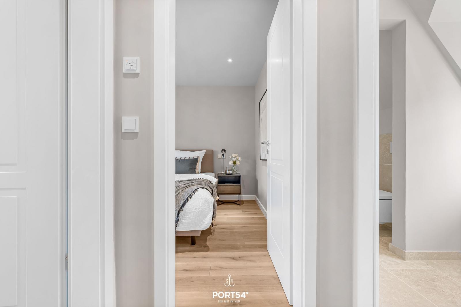 Schlafzimmer mit Bett, Nachttisch und Blumen. Holzboden und weiße Türen.