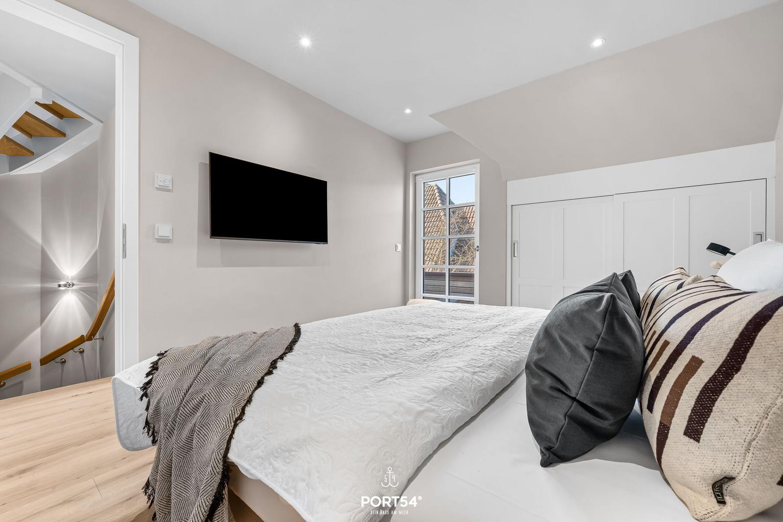 Schlafzimmer mit Bett, Fernseher und Fenster. Hinter dem Bett steht ein Schrank.