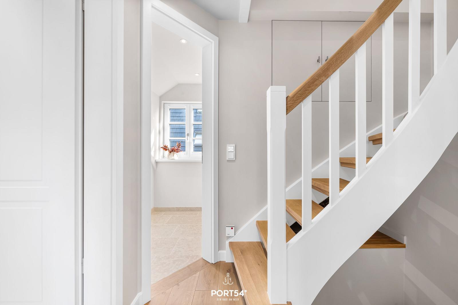 Treppenhaus mit Holzstufen und weißer Balustrade. Im Hintergrund ist ein Fenster mit Blumen zu sehen.