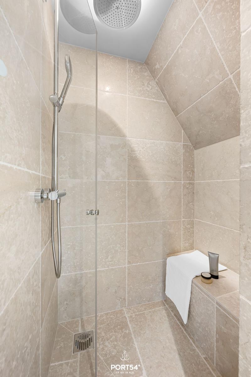 Dusche mit Duschbrause und Handbrause, hell verkleidet mit Steinplatten.