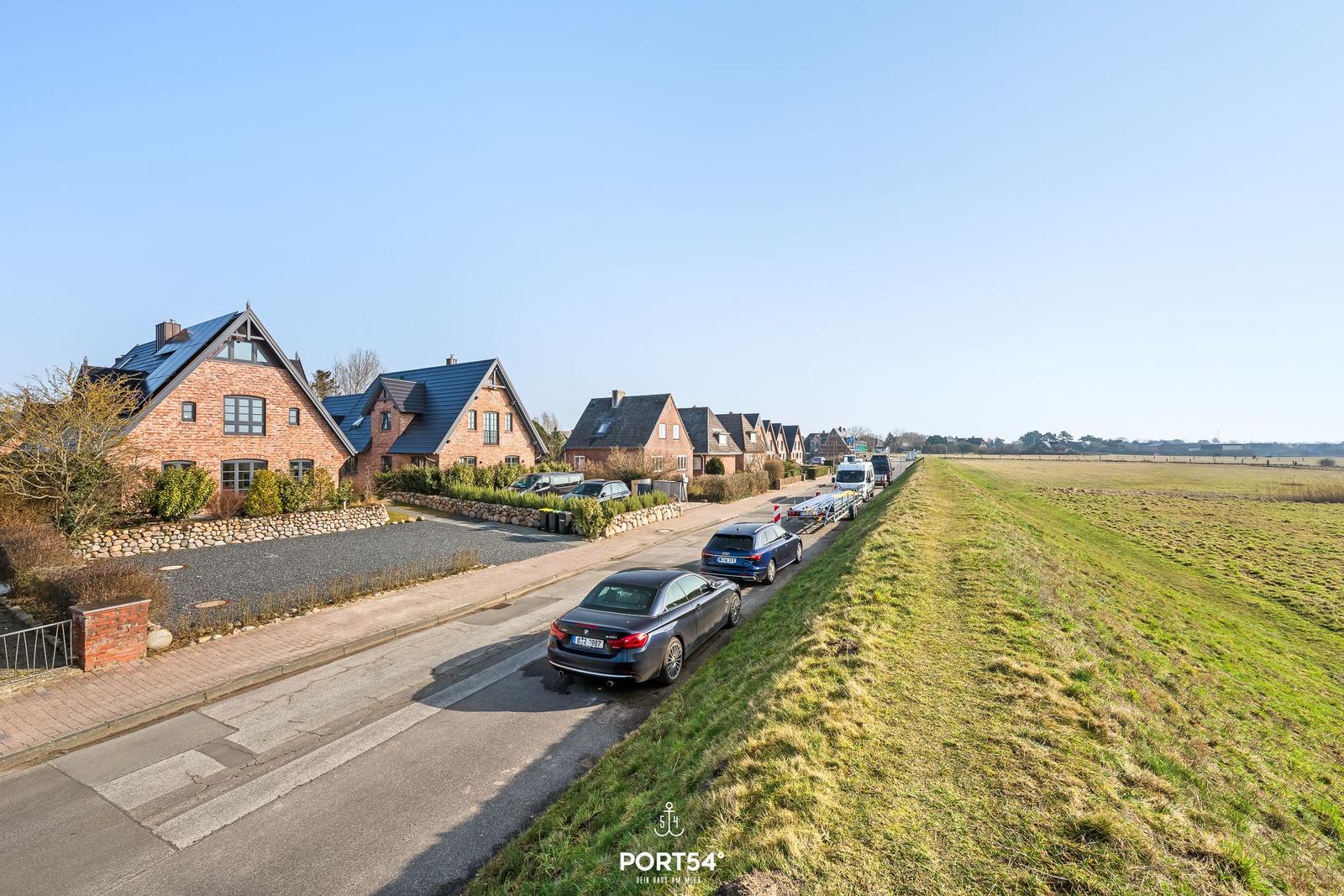 Reihe von Häusern mit dunklen Dächern entlang einer Straße mit Parkplätzen und grünem Feld.