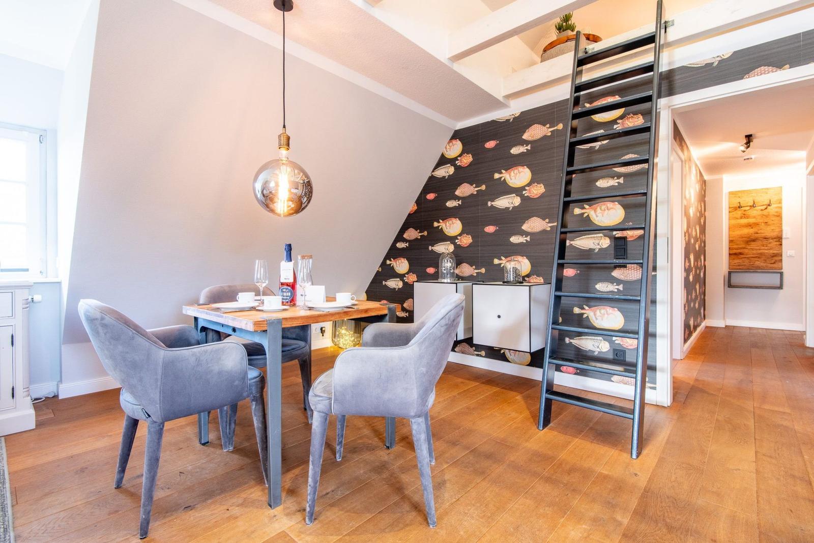 Essbereich mit Tisch, Stühlen und Wand mit Fischmotiv. Treppenhaus mit Leiter führt zur oberen Etage.