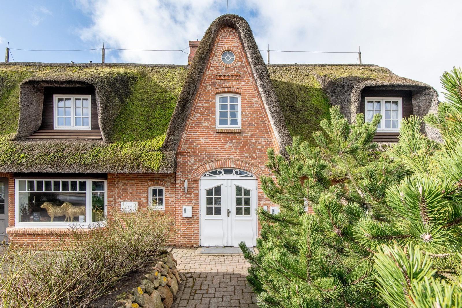 Haus mit strohgedecktem Dach, weiße Tür und Fenster, grüne Bäume im Vorgarten.