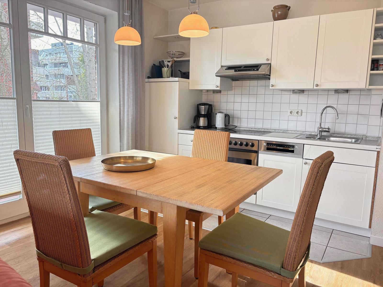 Essbereich mit Holztisch und Stühlen. Küche mit weißen Schränken und Arbeitsfläche.
