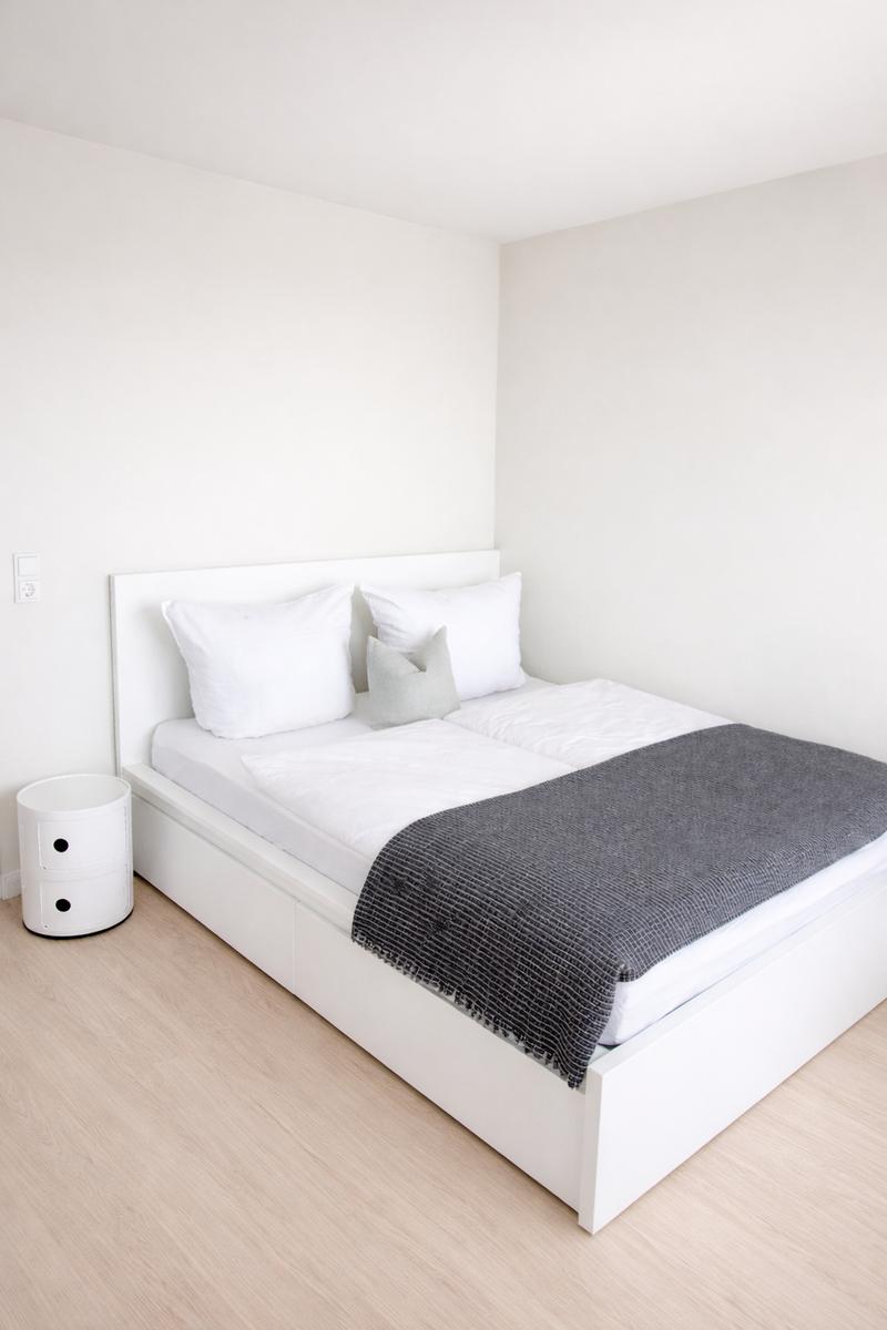 Schlafzimmer mit weißem Bett, grauer Decke und Nachttisch.