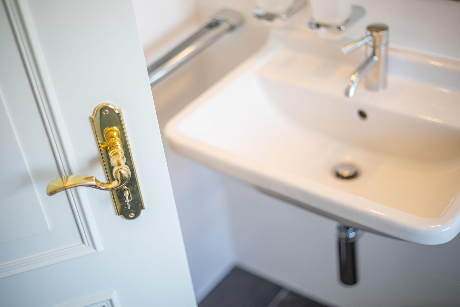 Hinter einer weißen Tür mit goldenem Griff ist ein Waschbecken mit Wasserhahn sichtbar.