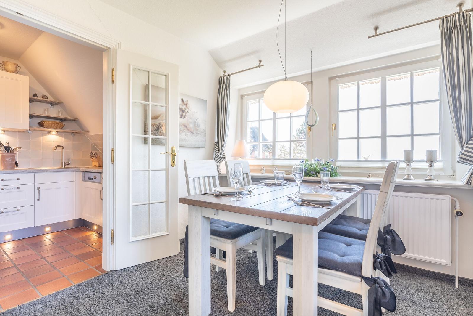 Esszimmer mit Tisch, Stühlen und Fenster. Küche im Hintergrund sichtbar.