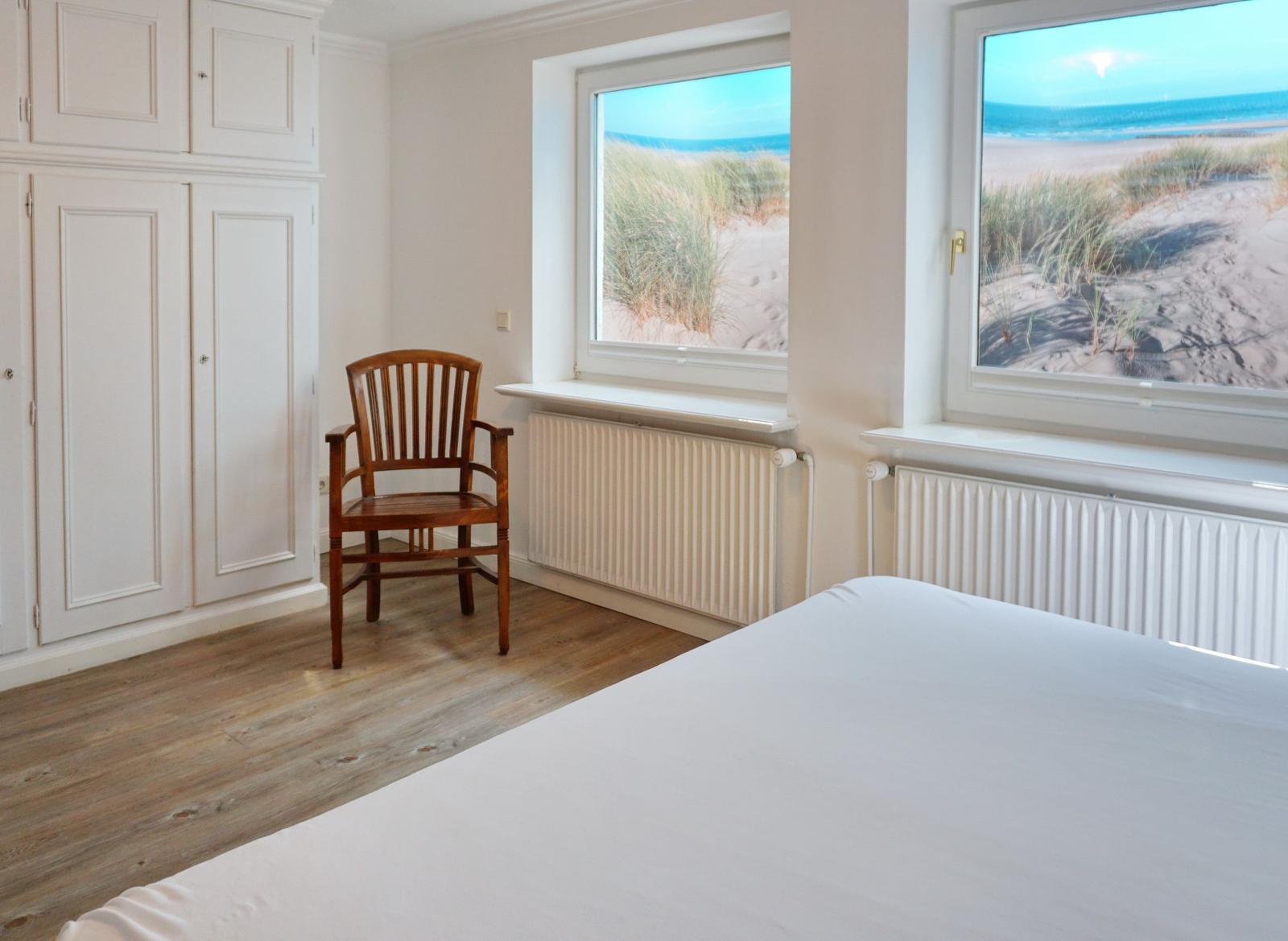 Schlafzimmer mit Blick auf Strand und Meer, weißes Bett, Holzstuhl und Kleiderschrank.