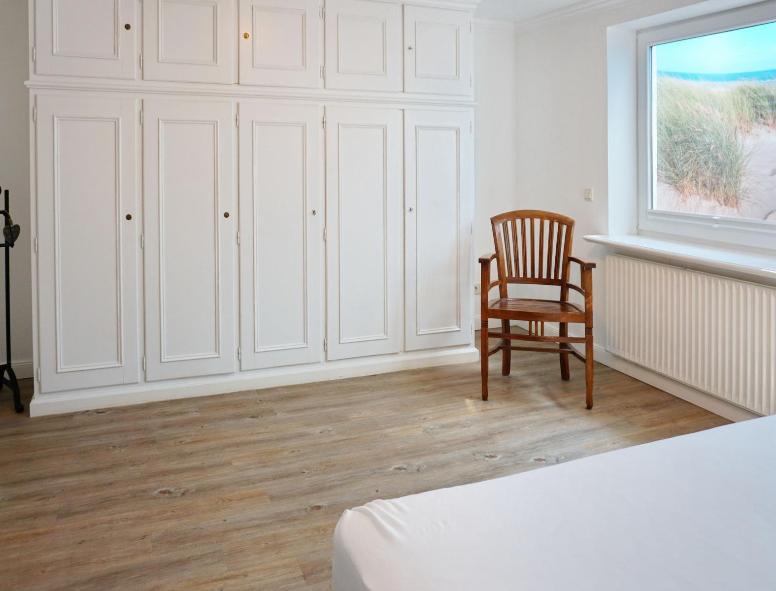Schlafzimmer mit weißem Schrank, Holzstuhl und Blick auf Strand.