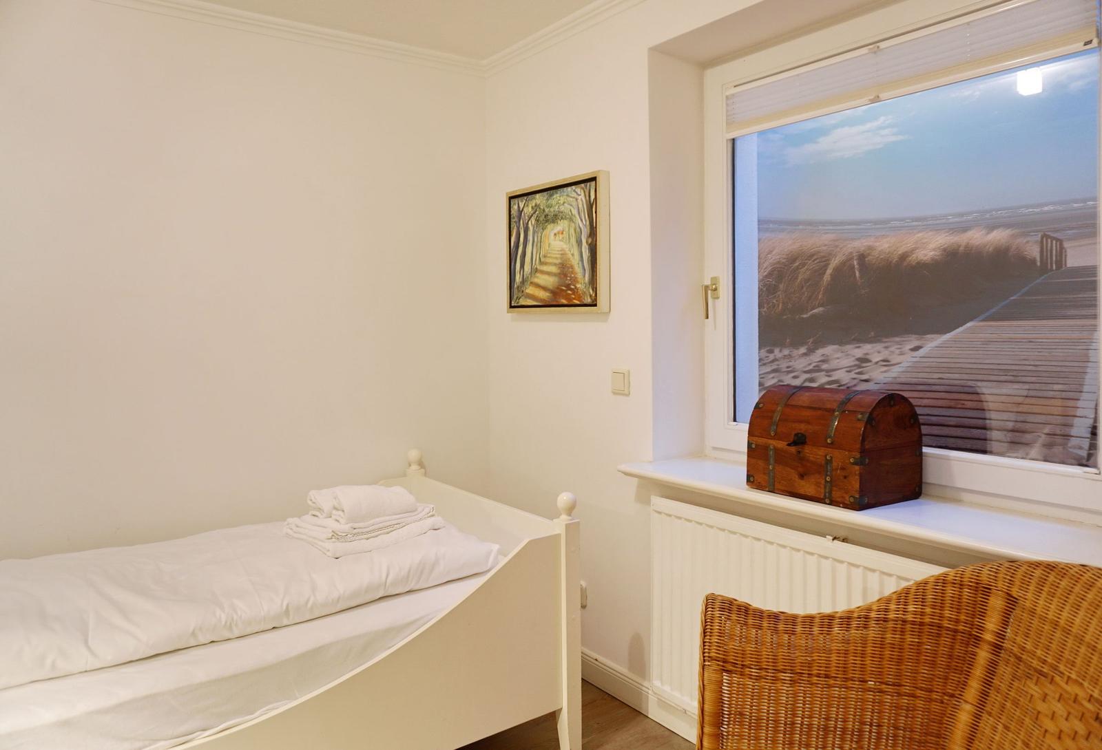 Ein Schlafzimmer mit Bett, Fenster und Kiste. Draußen Sanddünen und Holzweg.