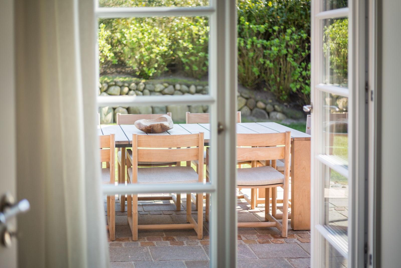 Durchgehende Glastüren führen auf eine Terrasse mit Holztisch und Stühlen. Hintergrund: grüne Hecke und Steinmauer.