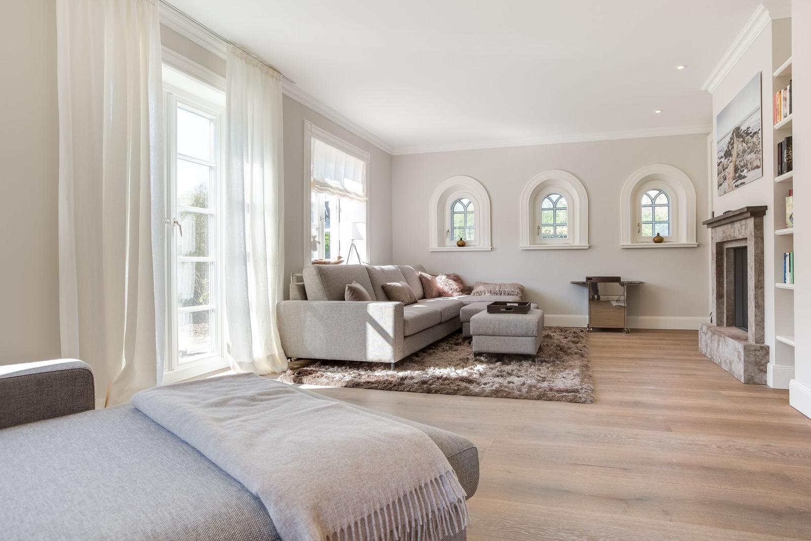 Wohnzimmer mit Sofa, Teppich und drei Bogenfenstern. Holzboden und Kamin sichtbar.