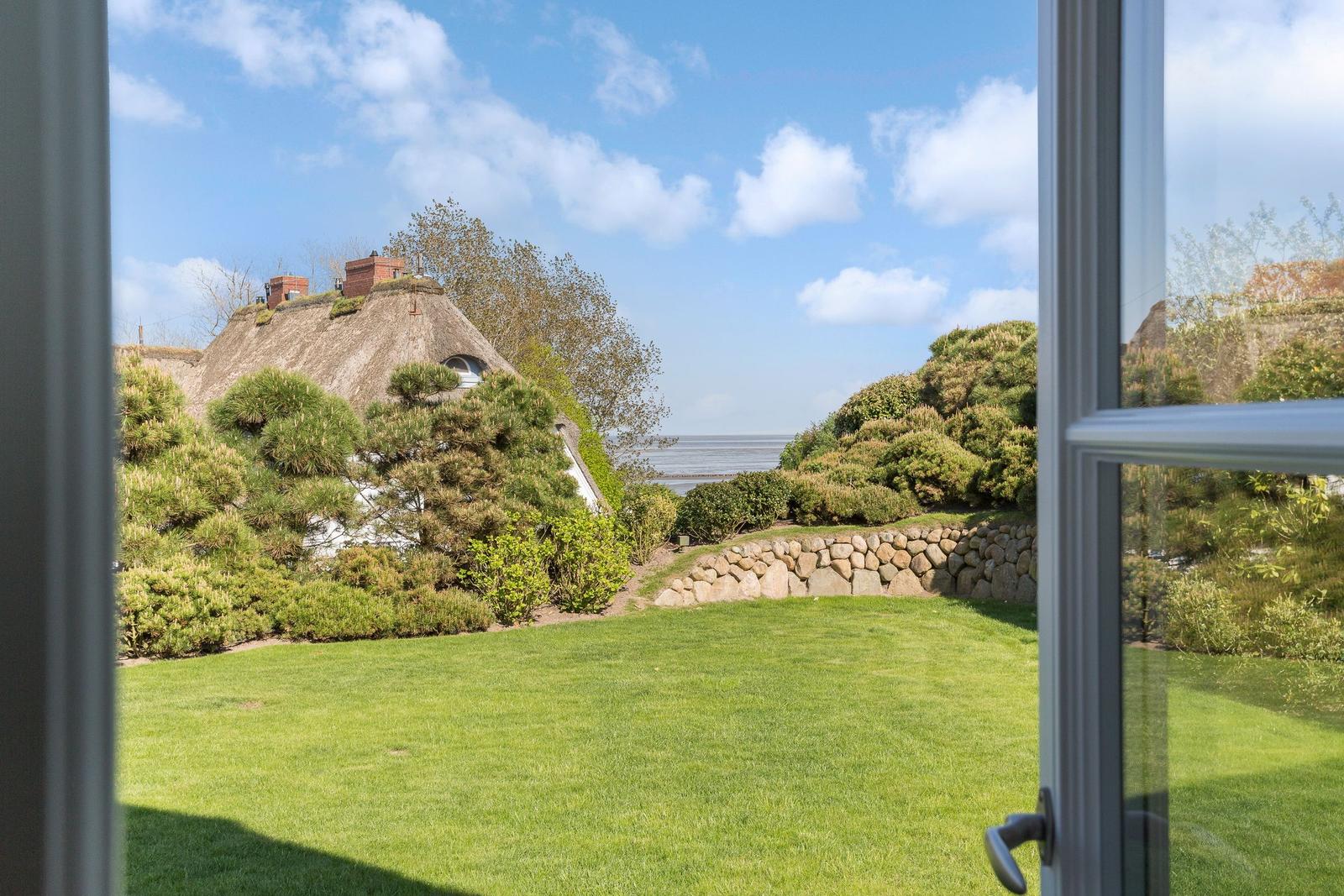 Von Fenster aus Blick auf grünen Garten, Steinmauer und traditionelles Haus mit Strohdach.