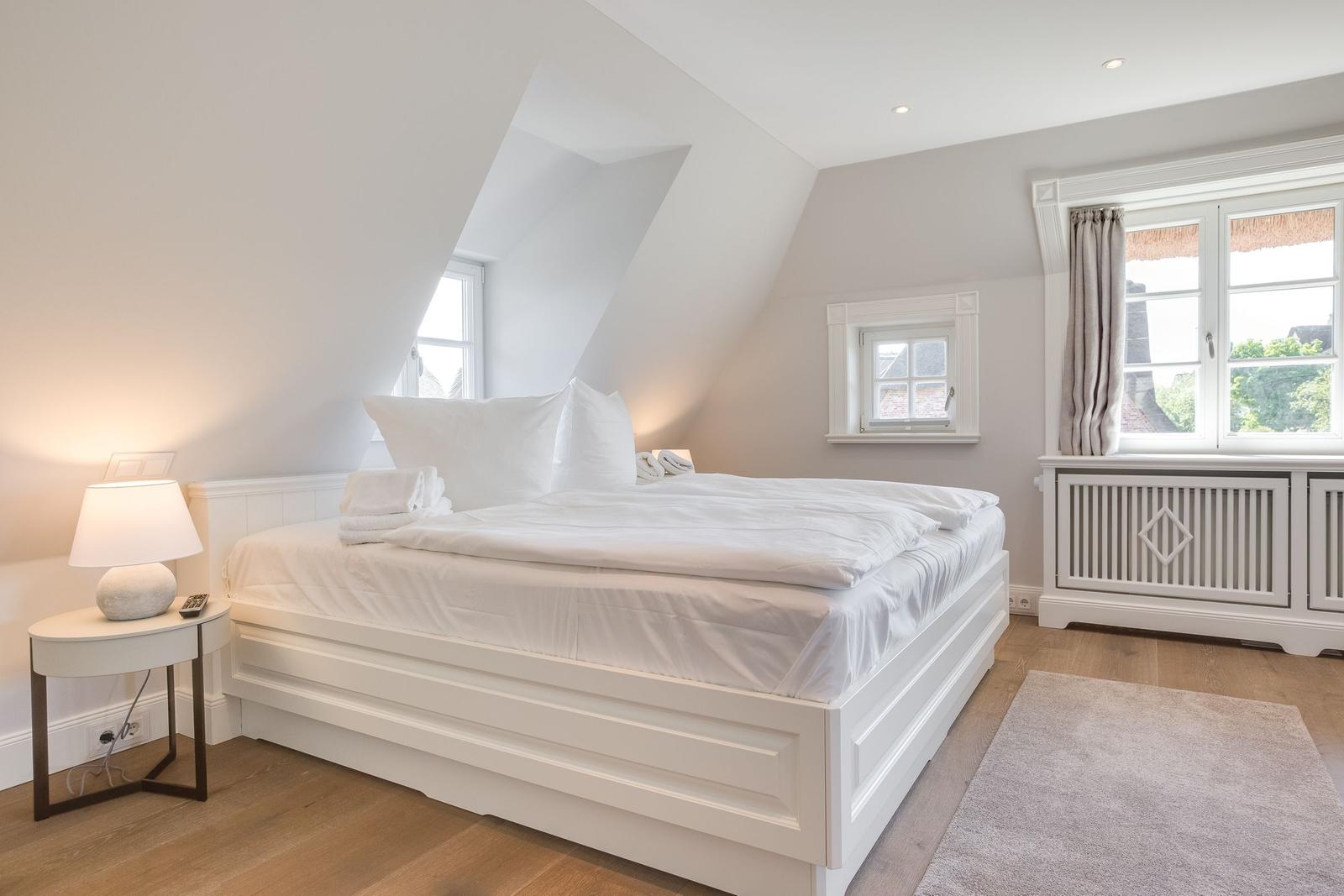 Schlafzimmer mit weißem Bett, Holzboden und Fenstern.