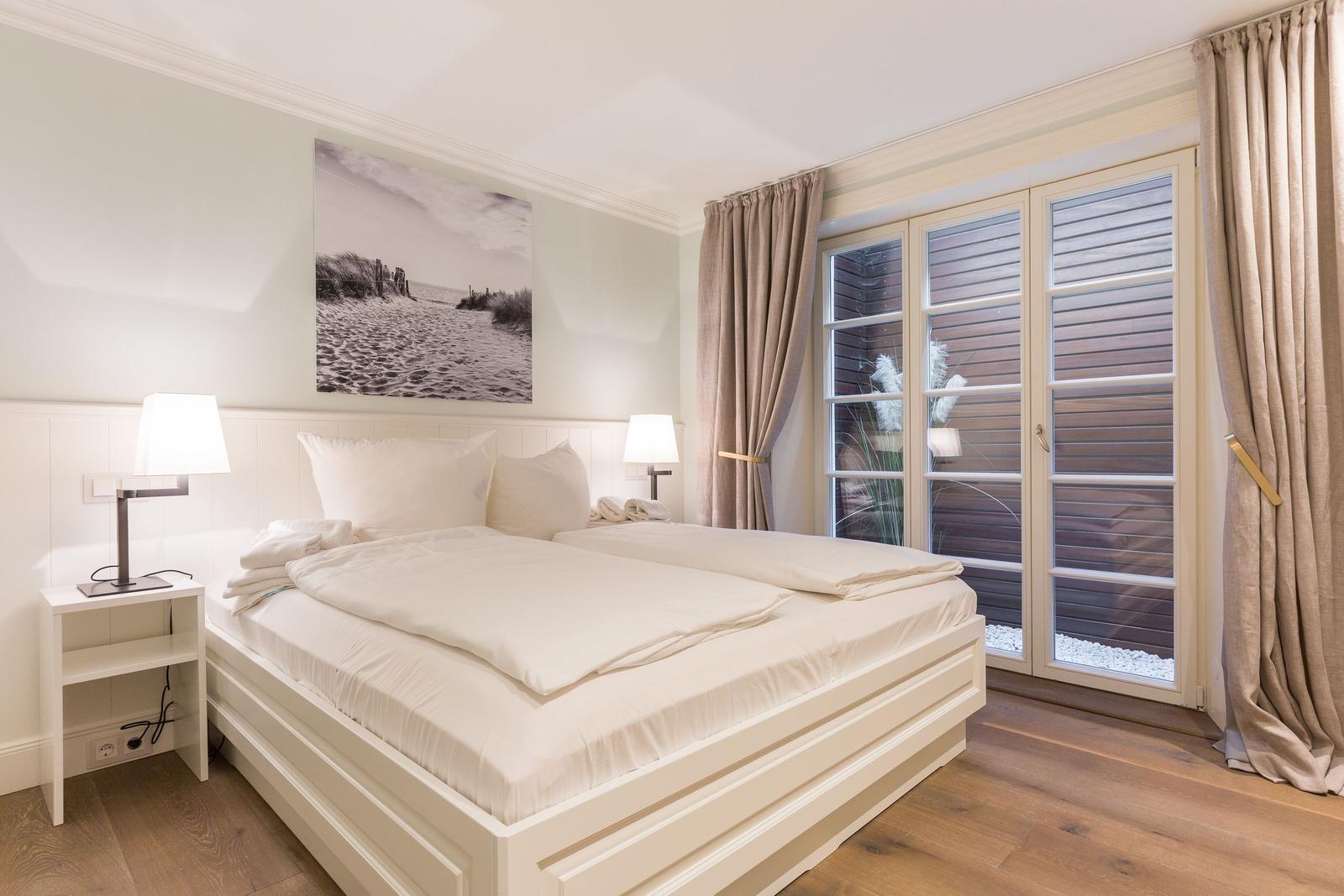 Ein Schlafzimmer mit weißem Bett, zwei Nachttischen und großen Fenstern.