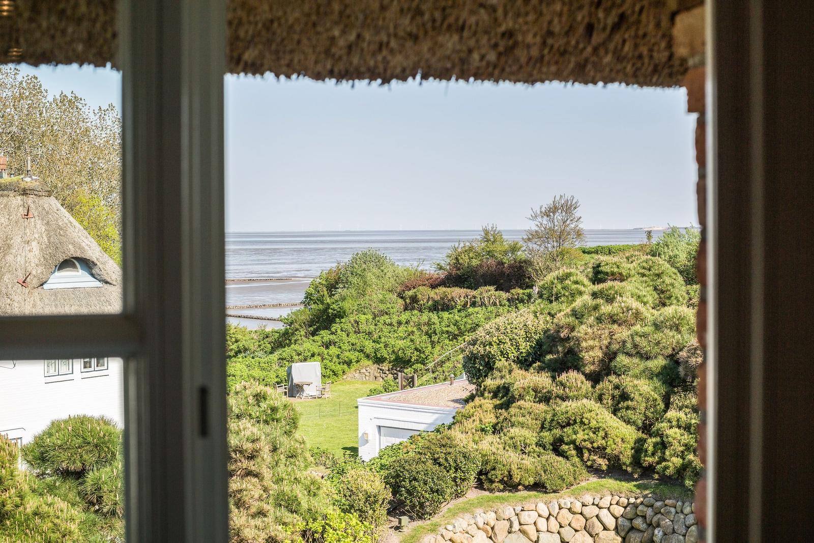 Durch das Fenster blickt man auf einen Garten mit Bäumen und das Meer.
