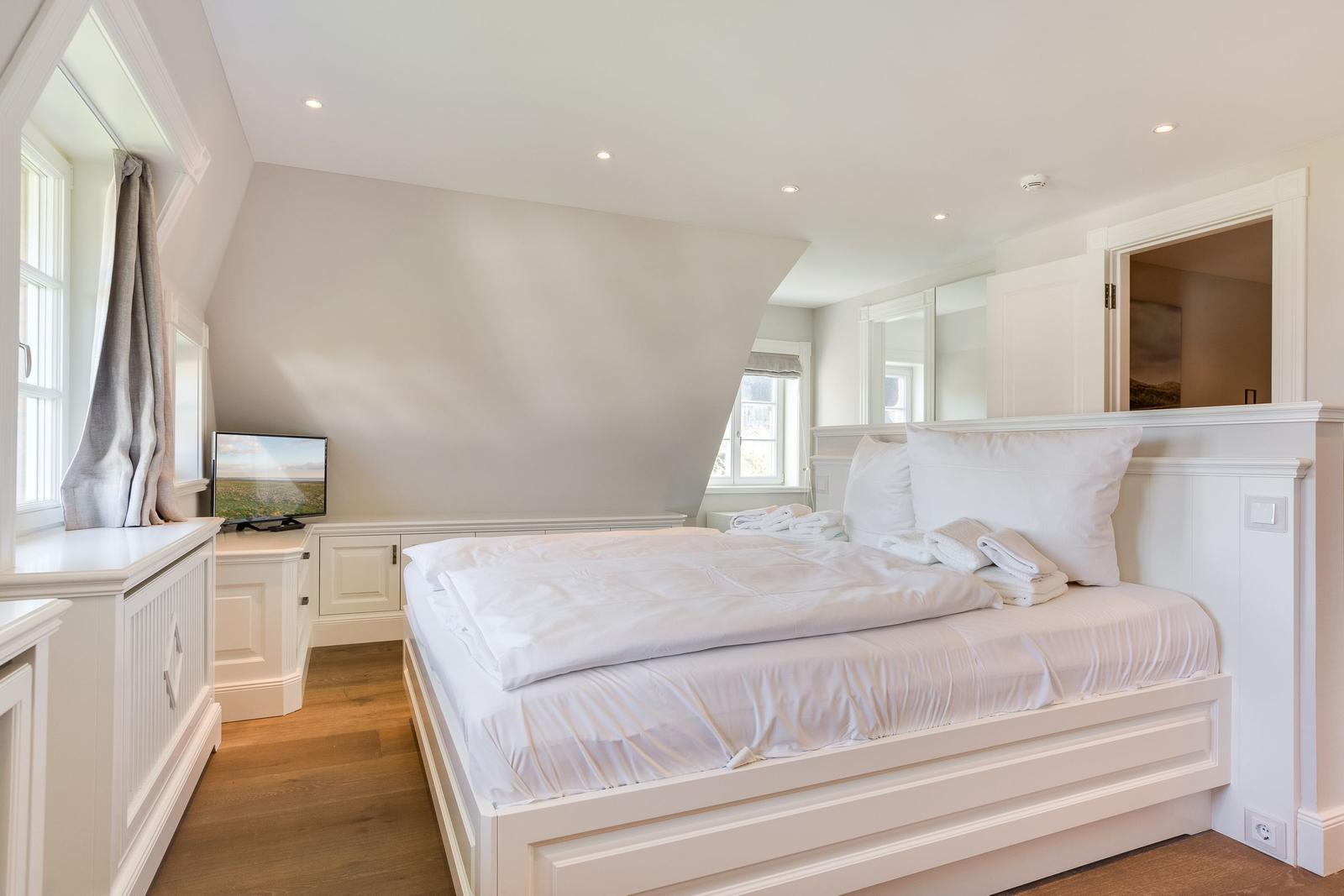 Schlafzimmer mit weißem Bett, TV und Holzboden. Dachfenster und Spiegel sorgen für Licht.
