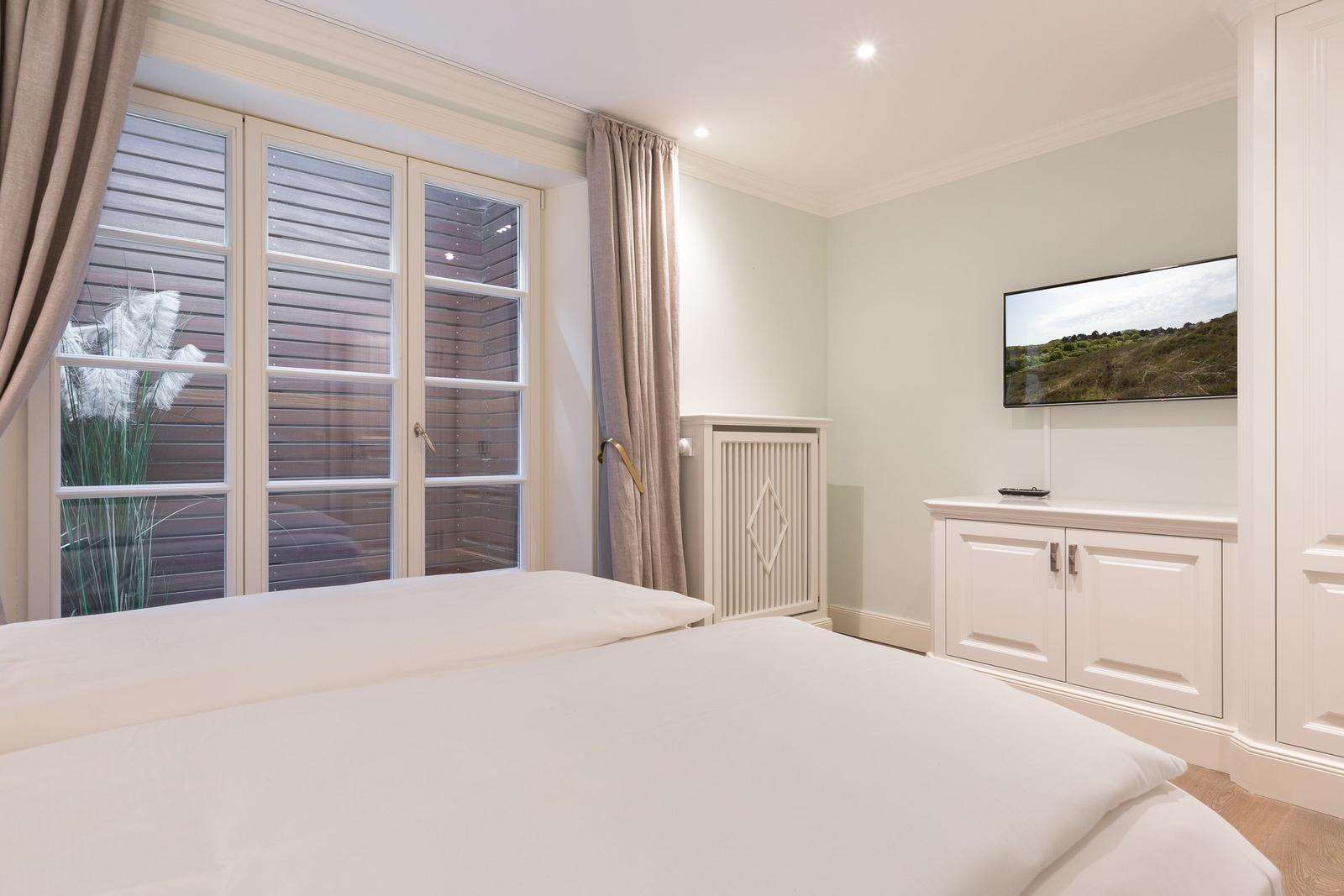 Schlafzimmer mit weißem Bett, Fenster und Fernseher an der Wand.