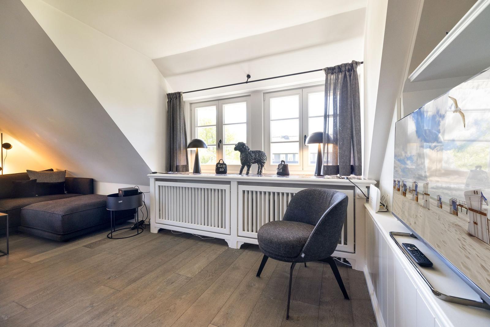 Ein Wohnzimmer mit Sofa, Stuhl und TV. Fenster mit Vorhängen und Heizkörper.