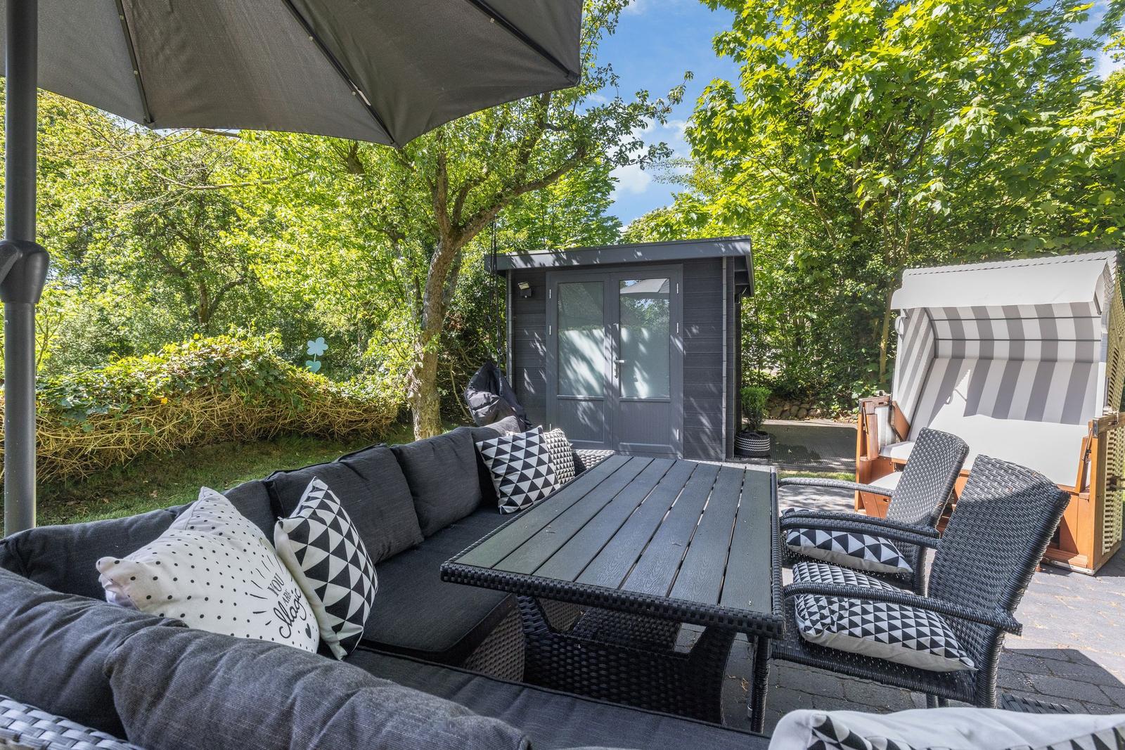 Terrasse mit Sitzgruppe, Tisch und Sonnenschutz. Hintergrund: Gartenhaus und Bäume.
