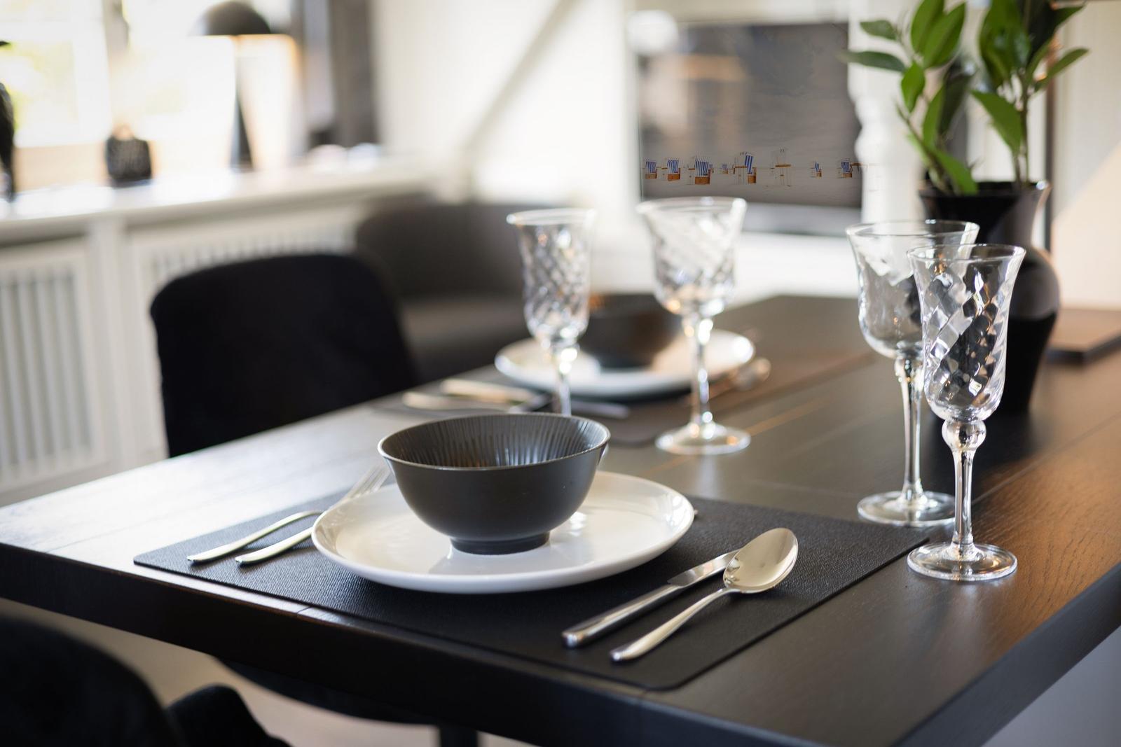 Esstisch mit Teller, Schale, Besteck und Gläsern. Hintergrund: Sofa und Pflanze.