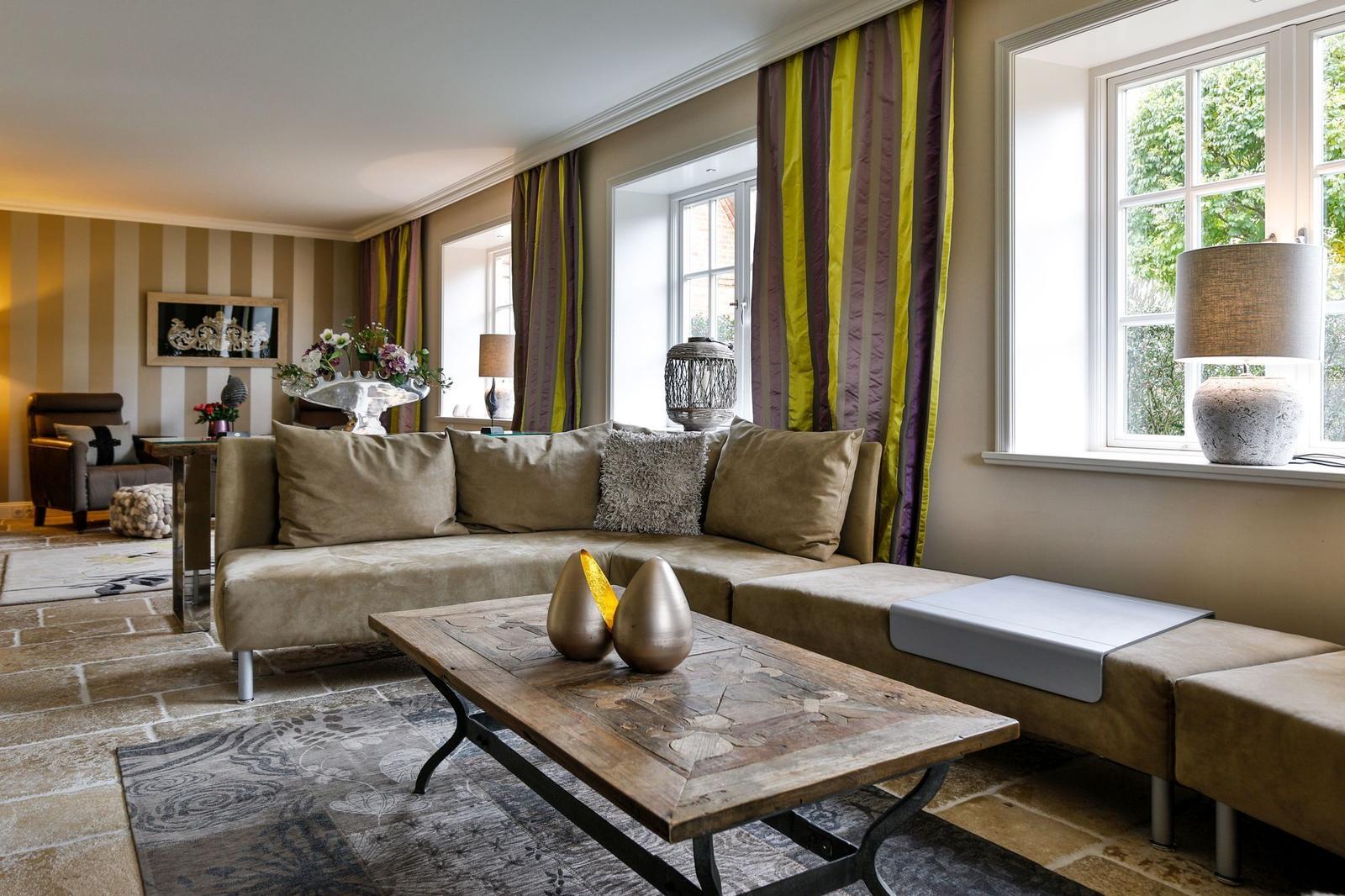 Ein Wohnzimmer mit Sofa, Tisch und Fenstern. Vorhänge und Wandstreifen sind sichtbar.