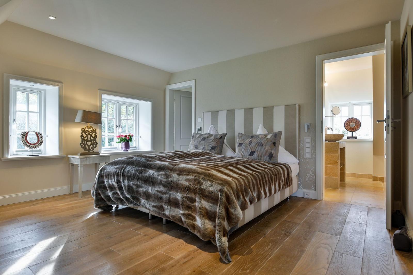 Schlafzimmer mit Bett, Holzboden und Fenstern. Blick in angrenzenden Raum.