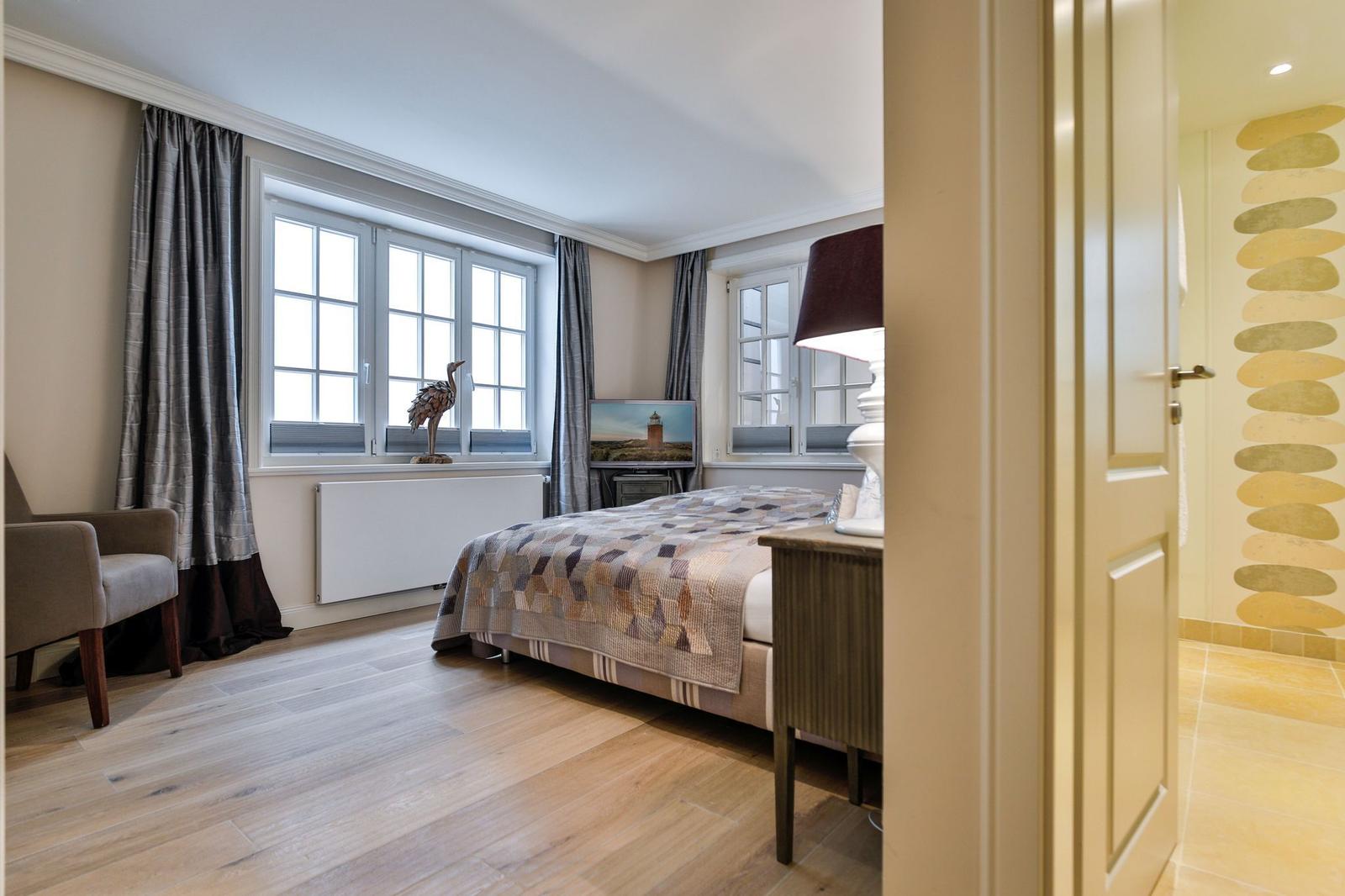 Schlafzimmer mit Bett, Fenstern und Holzboden. Tür führt in Bad.