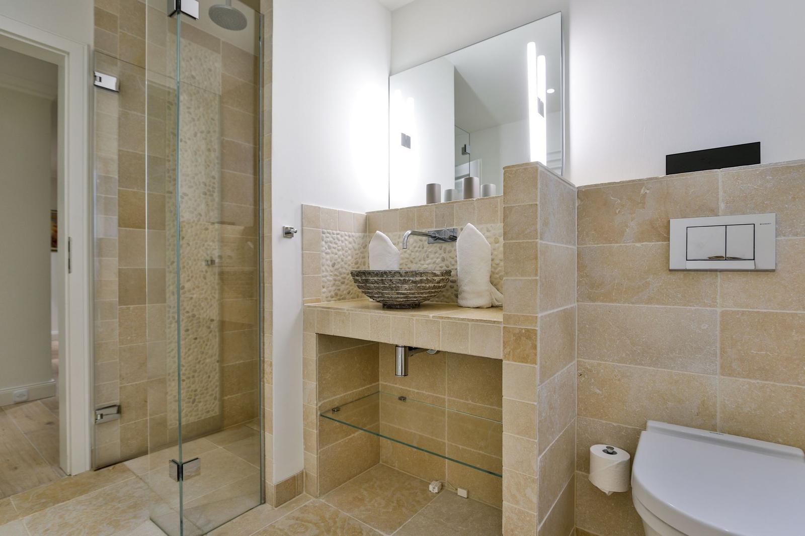 Badezimmer mit Dusche, Waschbecken und Toilette. Steinfliesen und Spiegelschrank.