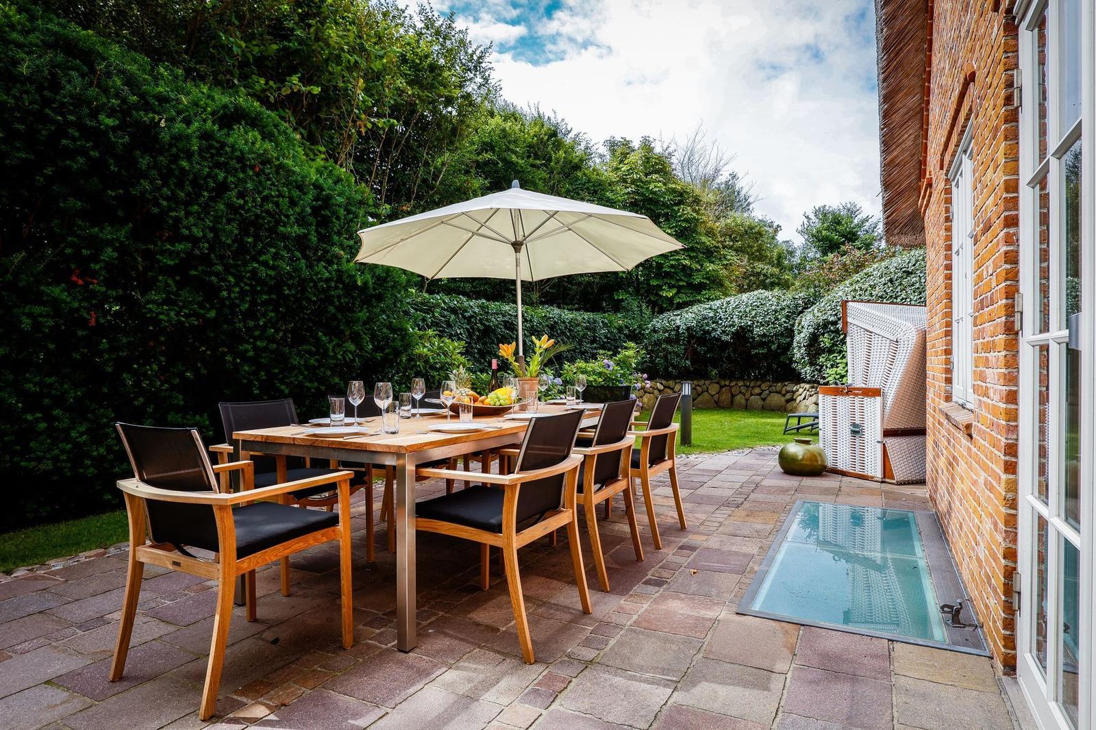Terrasse mit Tisch, Stühlen und Sonnenschirm. Garten mit Rasen und Hecken. Glasplatte neben Hauswand.