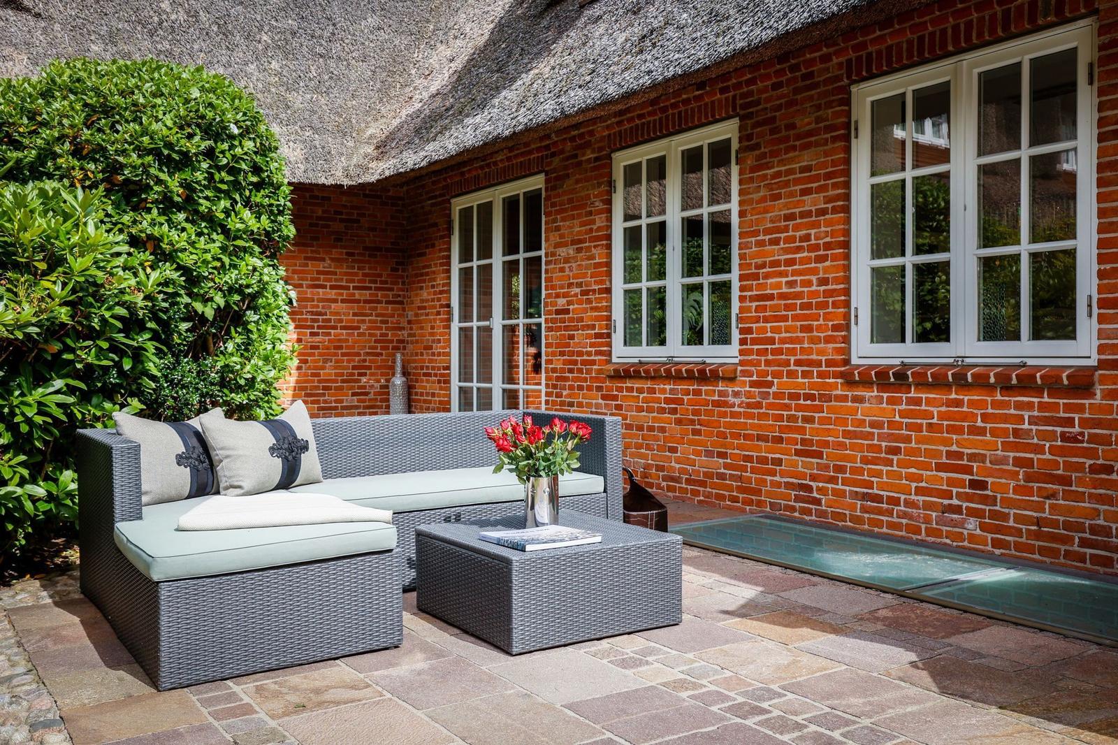 Terrasse mit Rattan-Sofa, Tisch und Blumen vor rotem Backsteinhaus mit Strohdach.