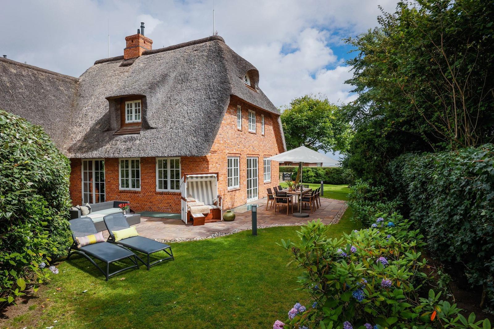 Haus mit Strohdach, Terrasse und Garten mit Liegen und Tisch.