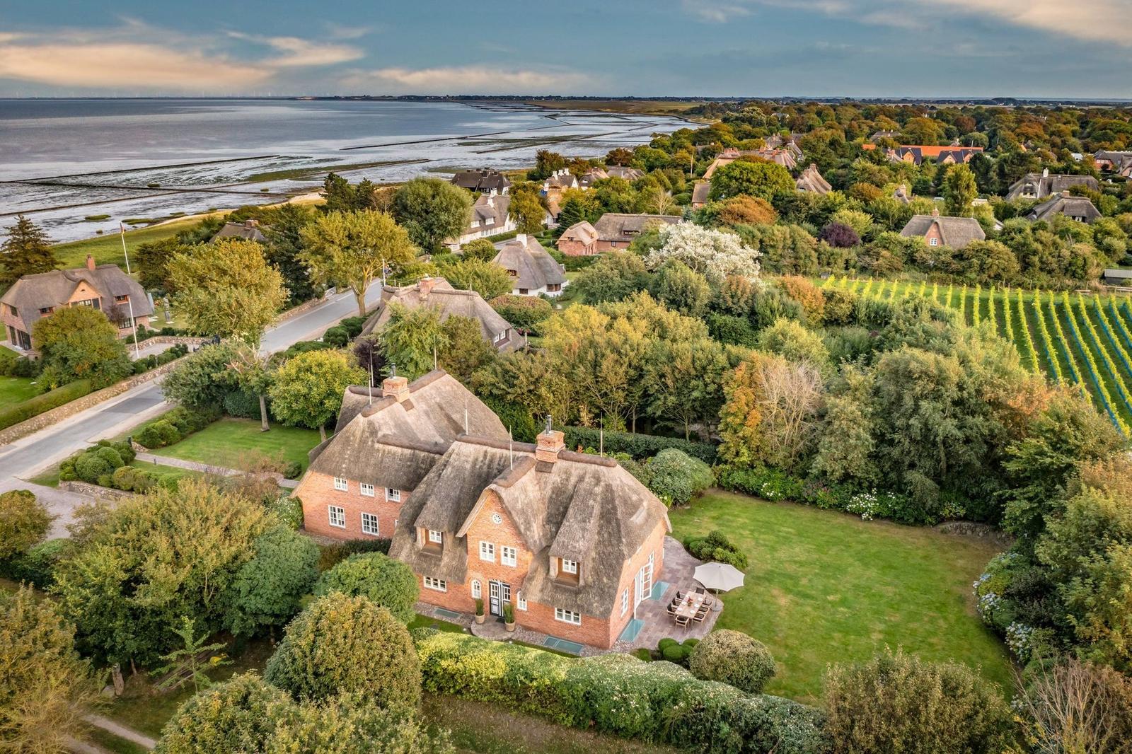 Traditionelles Haus mit Strohdach, umgeben von Garten und Wald. Blick auf Wasser und Landschaft.