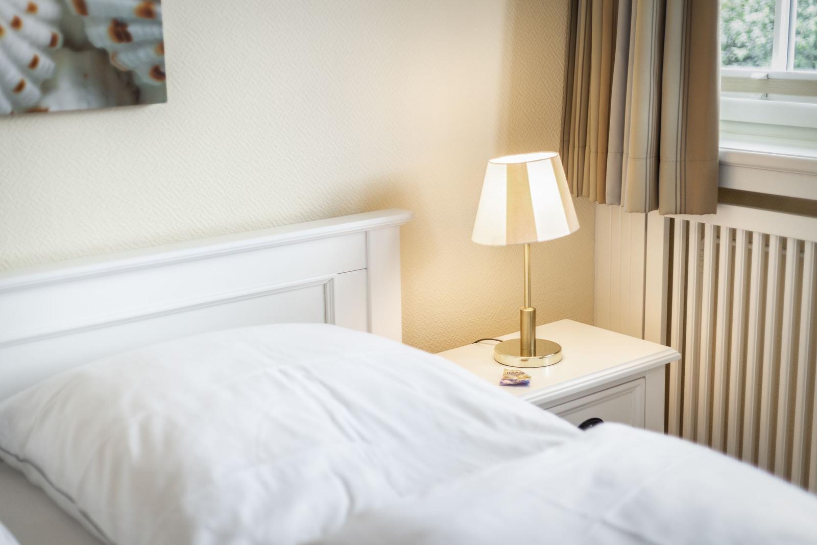 Schlafzimmer mit weißem Bett, Nachttisch und Lampe. Fenster mit Vorhängen.