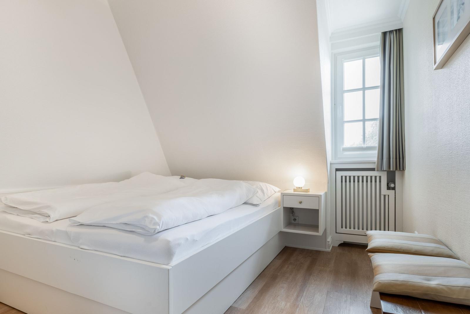 Schlafzimmer mit weißem Bett, Nachttisch und Fenster.