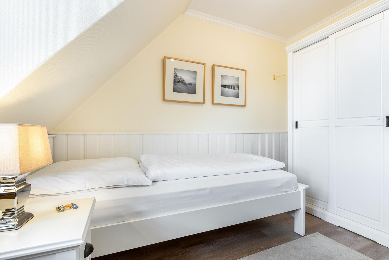 Schlafzimmer mit weißem Bett, Nachttisch und Kleiderschrank unter schräger Decke.