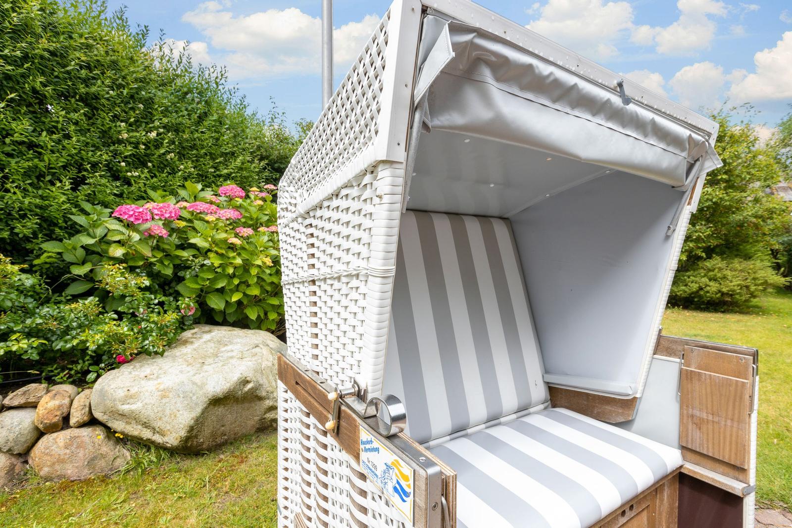 Weiße Strandkorb mit Streifenpolster im Garten, umgeben von Grün und Blumen.