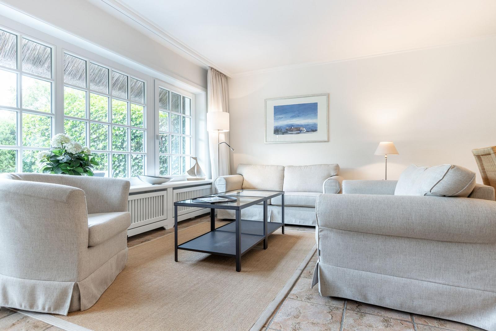 Wohnzimmer mit hellen Sofas, Glas-Tisch und großen Fenstern mit Blick auf Grün.