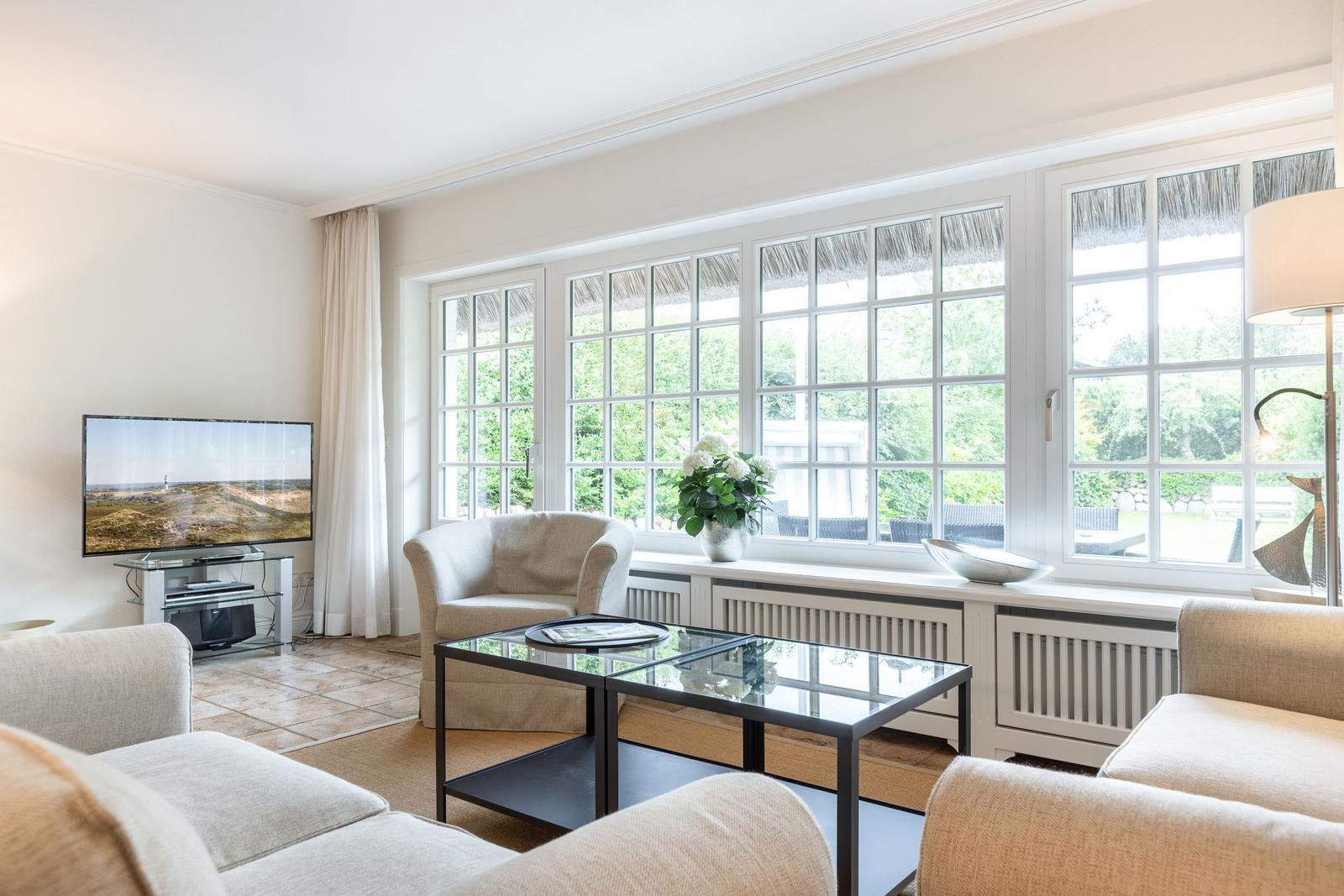 Wohnzimmer mit Sitzgelegenheiten, Fernseher und großem Fensterblick auf Garten.