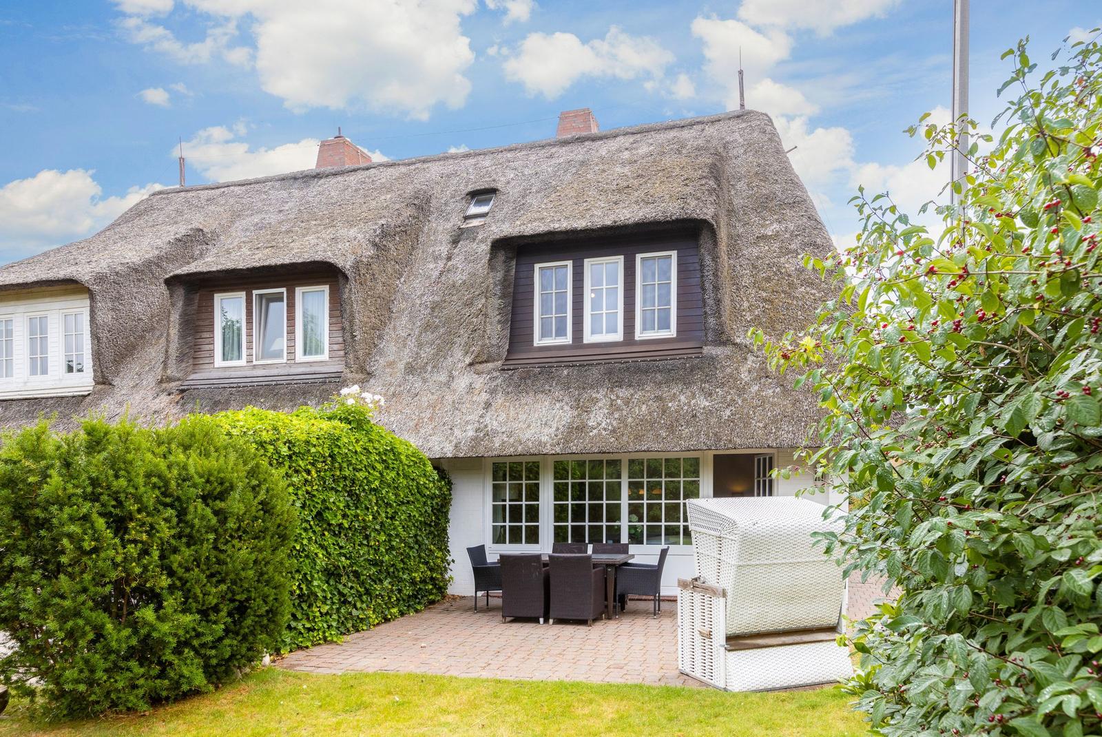 Haus mit Strohdach, Terrasse und Garten. Weiße Möbel und grüne Hecke.