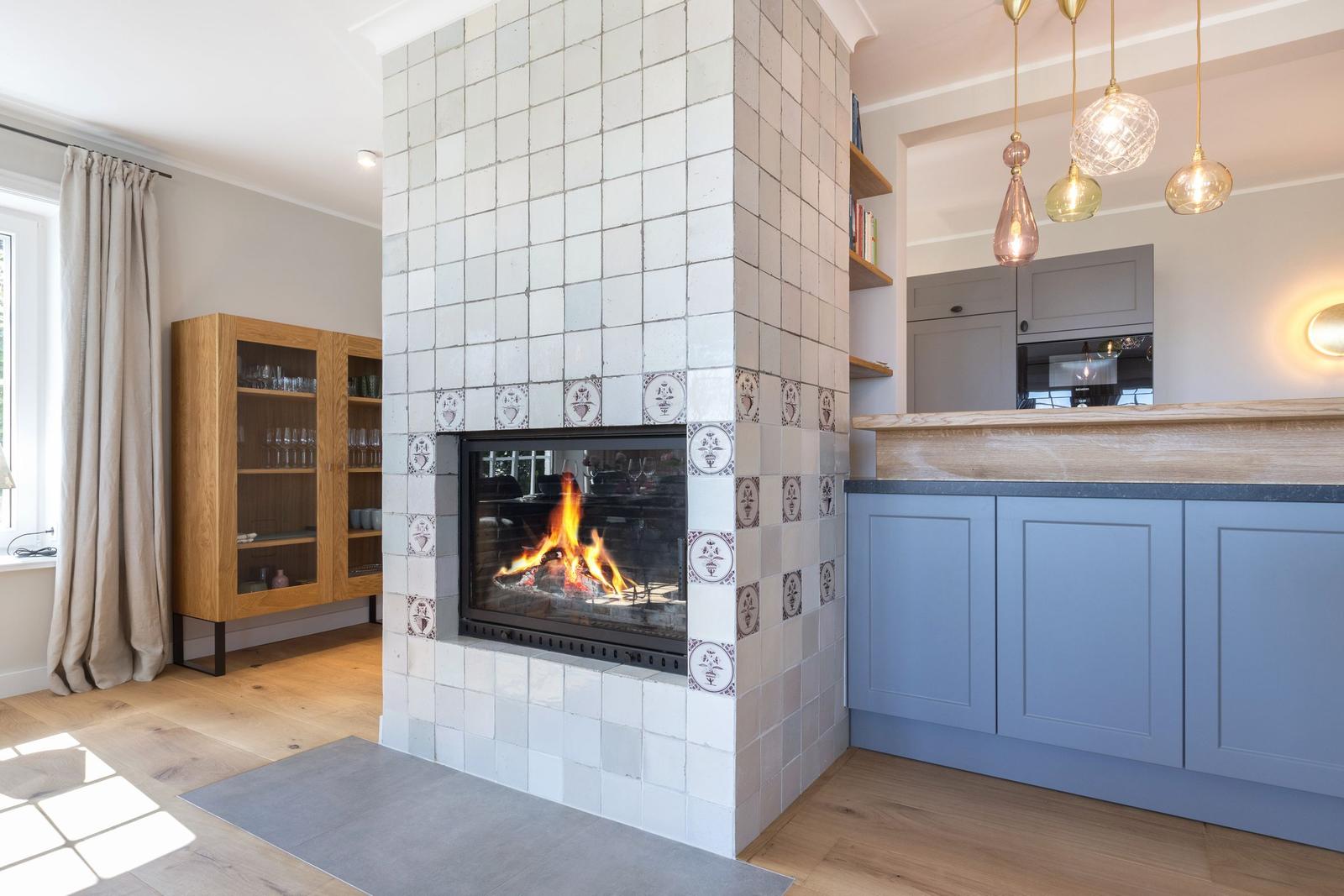 Kamin mit Flammen, Holzboden, Küchenzeile und hängende Lampen.