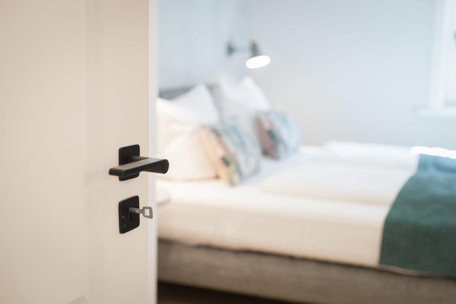Schlafzimmer mit Bett und Nachttischlampe. Tür mit schwarzer Klinke ist geöffnet.