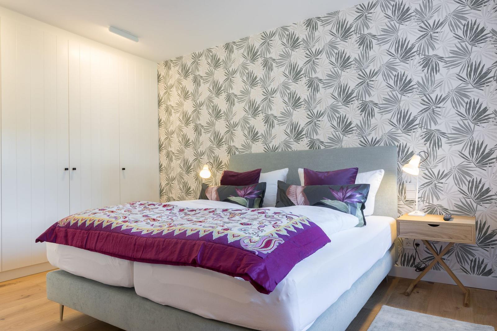 Schlafzimmer mit Bett, Nachttisch und Tapete mit Blattmuster.