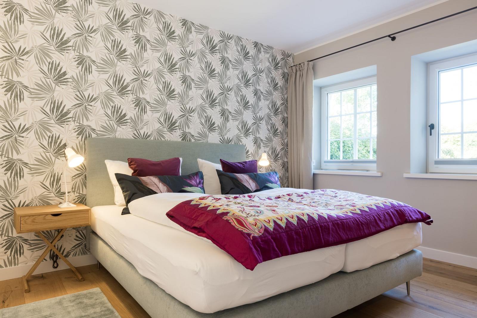 Schlafzimmer mit Bett, Kopfteil, Wandtapete und zwei Fenstern.