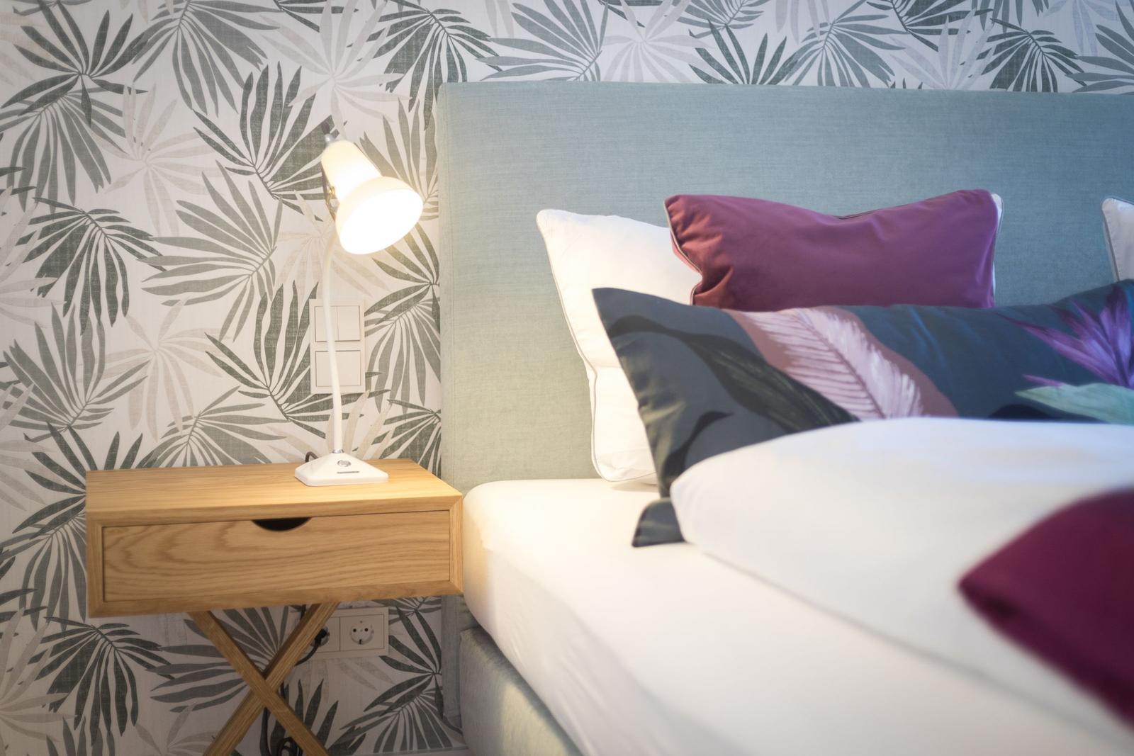 Schlafzimmer mit Bett, Nachttisch und Lampenwand mit Palmblattmuster.