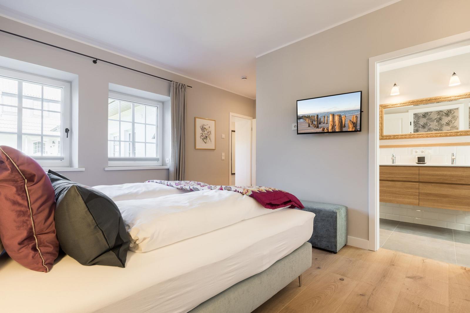 Schlafzimmer mit Bett, Fenstern und TV. Badezimmer im Anschluss.
